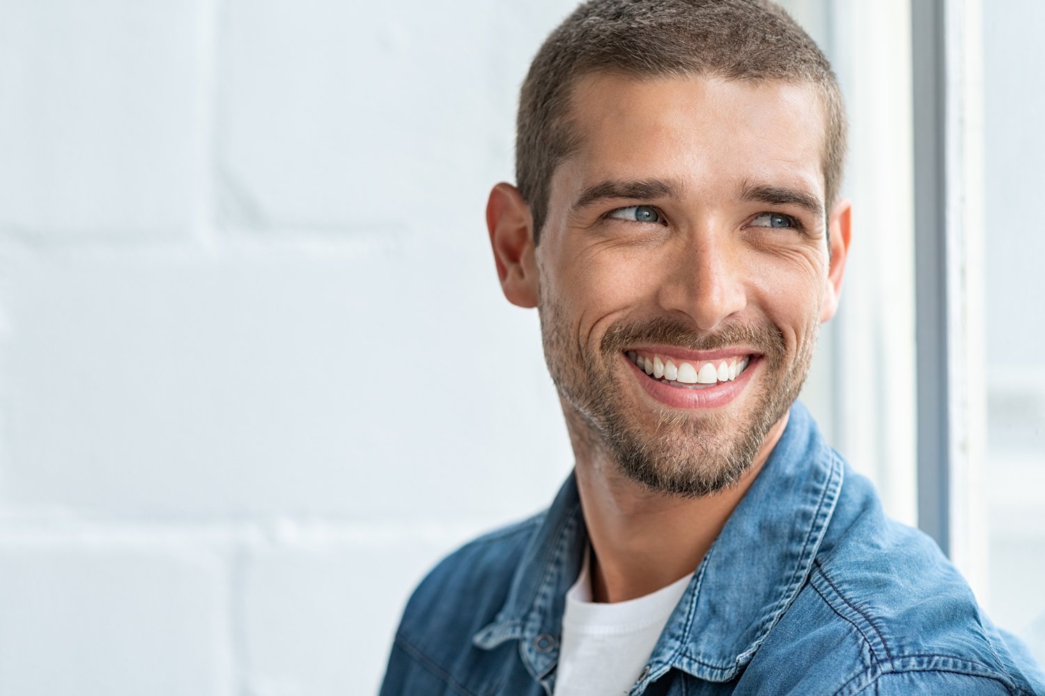A handsome man with short hair, blue eyes, and a beard smiles broadly, showing his white teeth as he looks off to the side. He is wearing a blue denim shirt against a bright, out-of-focus white background.