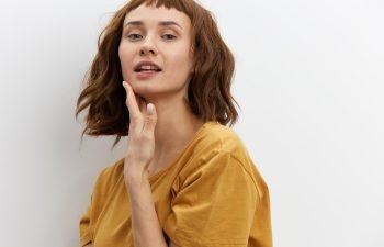 A young woman with a brown bob and bangs, wearing a yellow t-shirt, gently touches her jaw and neck while posing against a white wall.